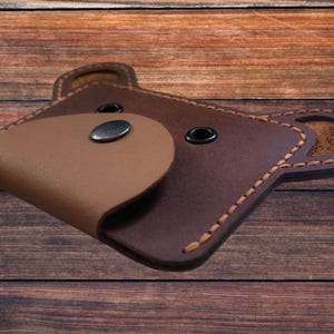 May include: A brown leather bear-shaped wallet with a light brown snout and black button eyes. The wallet has a snap closure and stitched edges. The design is a unique and playful accessory.