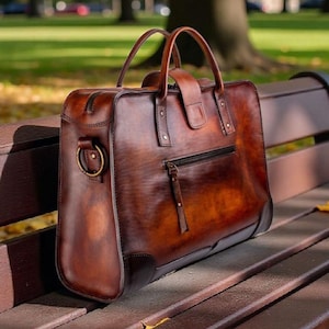 May include: A brown leather briefcase with a top handle, zipper closure, and a front pocket. The bag has a rectangular shape and is sitting on a wooden bench. The background shows a park with trees and green grass.