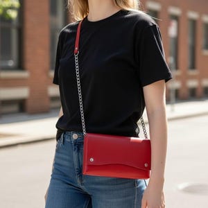 May include: A rectangular red leather crossbody bag featuring a smooth finish, a curved front flap with two silver-tone metal snap closures, and a thin silver-tone metal chain strap that rests against a black t-shirt and blue denim jeans.