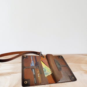 Op de afbeelding: Open bruin leren portemonnee met meerdere kaartsleuven, een paspoortvak en een polsbandje. De portemonnee is gevuld met kaarten en contant geld en heeft een donkerbruine rand met decoratieve stiksels.