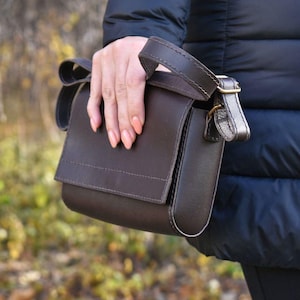 May include: A dark brown leather crossbody bag with a flap closure and adjustable strap. The bag has a rectangular shape with rounded edges and a gold-colored buckle. The bag is held by a person wearing a dark blue jacket.