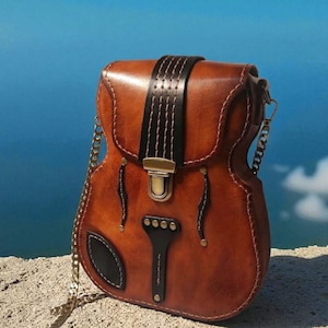 May include: A brown leather crossbody bag shaped like a guitar. The bag has a black strap and gold chain. The guitar-shaped bag has a black pickguard and gold hardware. The background is a blue sky.