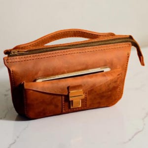 May include: A brown leather crossbody bag with a top handle and a front pocket. The bag has a zip closure and a gold-coloured clasp. A smartphone is visible in the front pocket. The bag is made of distressed leather.