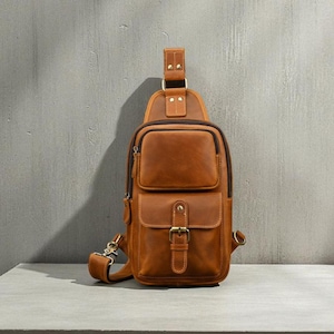 Puede incluir: Bolso bandolera de cuero marrón con múltiples bolsillos y cierre de hebilla. El bolso tiene un asa superior y una correa de hombro ajustable. El cuero tiene un aspecto envejecido y el bolso parece nuevo.