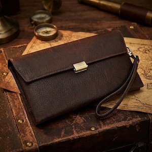 May include: A dark brown textured leather clutch wallet featuring a rectangular flap closure with a metallic silver-toned clasp and a matching leather wrist strap attached to the side. The wallet has a pebbled surface texture and a structured design.