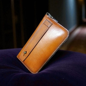 Puede incluir: Una cartera de cuero marrón con cierre de cremallera y cierre de botón a presión. La cartera es rectangular y tiene una superficie lisa y brillante. La cartera está sobre una superficie morada.