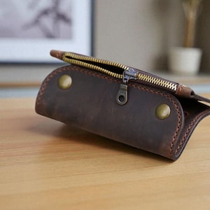 Op de afbeelding: Een bruine leren etui met een messing rits en twee messing klinknagels. De etui is gemaakt van distressed leer en heeft een gebogen vorm. De rits is open en onthult de binnenkant. De etui ligt op een houten oppervlak.