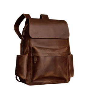 Puede incluir: Una mochila de cuero marrón con un bolsillo frontal grande y dos bolsillos laterales. La mochila tiene un cierre de solapa y correas ajustables.