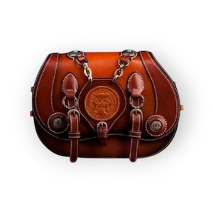 May include: A brown leather saddlebag with decorative stitching and silver-toned hardware. The bag features a central embossed emblem and turquoise accents. The design includes two buckle closures and a chain strap.