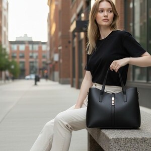 May include: A black leather tote bag featuring a structured silhouette, dual top handles, and a central vertical strap detail with a metallic buckle closure. The bag has a smooth finish and is designed with a wide, open-top opening for storage.