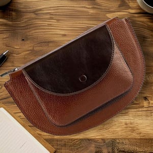 May include: A brown leather fanny pack with a dark brown flap and a zipper closure. The bag has a textured surface and a curved shape. The fanny pack is on a wooden surface.