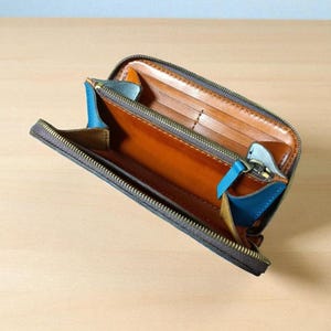 May include: Open, brown leather wallet with a zipper closure. The interior features multiple card slots and a contrasting blue accent. The wallet is set against a light wooden surface.
