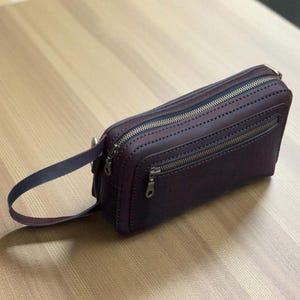 May include: A burgundy leather clutch with a handle. The rectangular bag has two zippered compartments and a smaller zippered pocket. The bag has decorative stitching and gold-colored zippers.