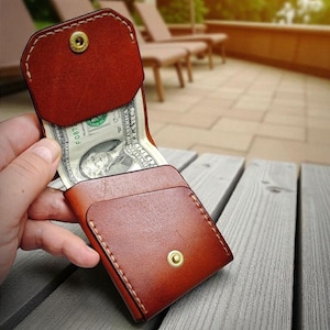 Op de afbeelding: Een bruine leren portemonnee met een drukknoopsluiting, met een Amerikaanse dollarbiljet erin. De portemonnee is open en onthult het geld binnenin. De portemonnee ligt op een houten oppervlak.