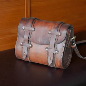 Könnte beinhalten: Braune Leder-Umhängetasche mit Klappenverschluss und verstellbarem Riemen. Die Tasche hat zwei vertikale Riemen mit Schnallen auf der Vorderseite und ein kleines geprägtes Design auf der Klappe. Die Tasche ist ca. 25 cm breit.