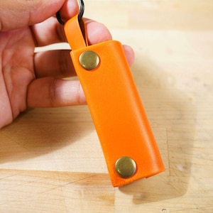 May include: Orange leather key holder with brass-colored snap closures and a black key ring. The rectangular case is made of smooth leather and has a loop for attaching to a belt or bag. A simple and functional accessory.