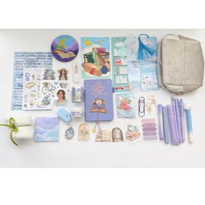 May include: A collection of stationery items, including a purple journal, stickers, pens, and washi tape. The items are arranged on a white surface. The journal has the text "Between Pages & Wild Moon". The color palette is soft, with shades of purple, blue, and white.