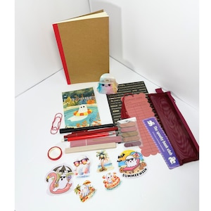 May include: A stationery set including a tan notebook with a red spine, a ghost-shaped resin figure, a postcard with a ghost in a pool, a red paperclip, pens, a red mesh pencil case, a library card bookmark, and several ghost-themed stickers.