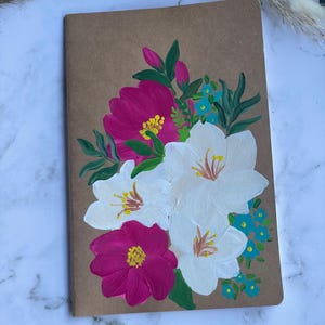May include: A hand-painted floral design on a brown paper notebook cover. The design features white, pink, and blue flowers with green leaves.