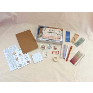 Könnte beinhalten: Ein Schreibwaren-Set in einer weißen Schachtel mit der Aufschrift Mini Stationery Kit, bestehend aus einem braunen Notizbuch, einem Checklisten-Block, verschiedenen buchthematischen Aufklebern, drei Stiften in Helllila, Weiß und Orange, Klebeband.