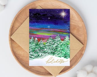May include: A holiday greeting card featuring a winter landscape with snow-covered evergreen trees under a starry night sky. The card has a gold script that says "happy holidays". The card is inside a brown envelope and placed on a wooden plate.