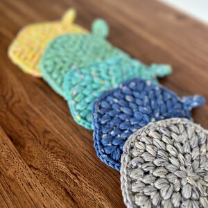 May include: Five crocheted pot holders in a rainbow of colors, from yellow to gray, arranged in a row on a wooden surface.