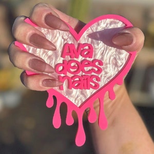 Pode incluir: Design de gotejamento rosa em forma de cora&ccedil;&atilde;o com o texto "Ava does nails" em letras brancas. O cora&ccedil;&atilde;o &eacute; segurado por uma m&atilde;o com unhas marrons escuras.
