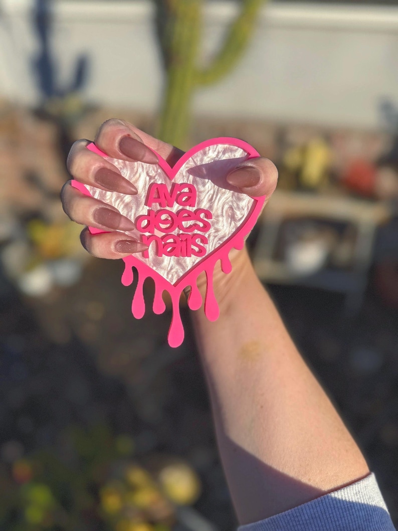 Pode incluir: Design de gotejamento rosa em forma de cora&ccedil;&atilde;o com o texto "Ava does nails" em letras brancas. O cora&ccedil;&atilde;o &eacute; segurado por uma m&atilde;o com unhas marrons escuras.