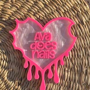 Pode incluir: Uma bandeja de resina em forma de cora&ccedil;&atilde;o rosa com um design de m&aacute;rmore branco e o texto "Ava does nails" em letras rosa. A bandeja tem um efeito de gotejamento na parte inferior.