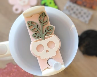May include: A pink and white skull-shaped cup holder with a green plant design. The holder is designed to fit around a straw and keep it in place.
