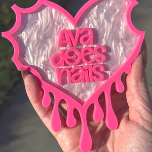 Pode incluir: Porta-copos de resina em forma de cora&ccedil;&atilde;o rosa com um centro branco iridescente e as palavras "Ava does nails" em letras rosa. O porta-copos tem um efeito de gotejamento ao redor das bordas.