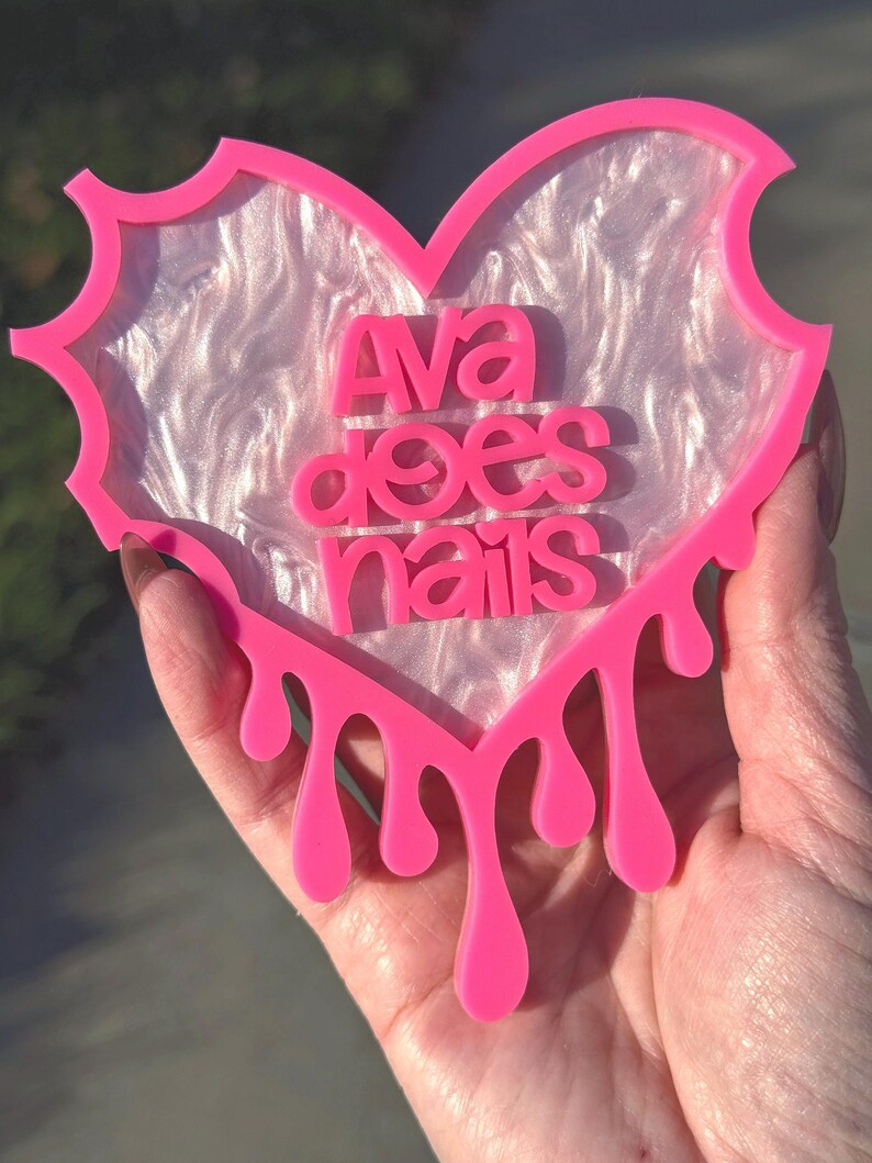 Pode incluir: Porta-copos de resina em forma de cora&ccedil;&atilde;o rosa com um centro branco iridescente e as palavras "Ava does nails" em letras rosa. O porta-copos tem um efeito de gotejamento ao redor das bordas.