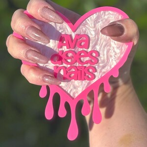 Pode incluir: Um letreiro de acr&iacute;lico em forma de cora&ccedil;&atilde;o rosa com bordas pingando. O letreiro diz "Ava does nails" em letras brancas sobre um fundo marmorizado branco e rosa. Uma m&atilde;o com unhas compridas, rosadas e brilhantes est&aacute; segurando o letreiro.