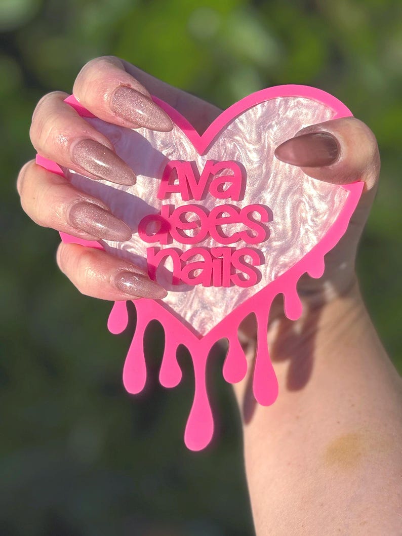 Pode incluir: Um letreiro de acr&iacute;lico em forma de cora&ccedil;&atilde;o rosa com bordas pingando. O letreiro diz "Ava does nails" em letras brancas sobre um fundo marmorizado branco e rosa. Uma m&atilde;o com unhas compridas, rosadas e brilhantes est&aacute; segurando o letreiro.