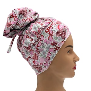 May include: A pink and white patterned scrub cap featuring hearts, mushrooms, butterflies, and peace signs. The cap is tied in a bun on top of the head. The fabric has a floral design with red and pink accents.