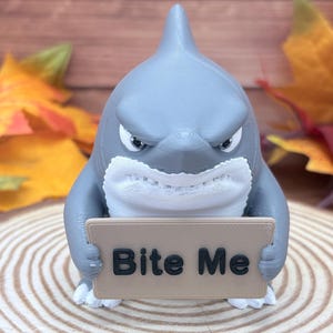 May include: A gray shark figurine with a white belly and fins, featuring a stern facial expression. The shark holds a rectangular tan sign in its front fins with the text "Bite Me" printed in bold black letters on the front of the sign.