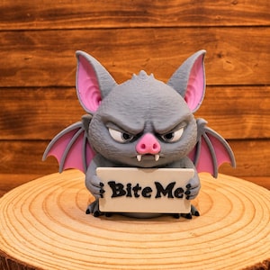 May include: A gray bat figurine with large pointed ears, pink inner ear details, and pink wings. The bat has a grumpy facial expression with two white fangs and holds a white rectangular sign with the black text "Bite Me" in the center.