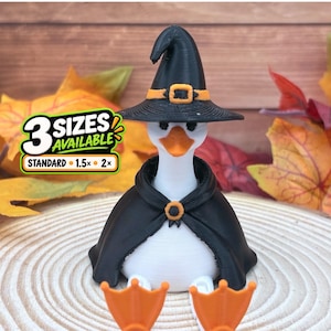 May include: A white goose figurine wearing a black witch hat with an orange buckle and a black cape with an orange clasp. The goose has orange webbed feet. Text on the image reads 3 SIZES AVAILABLE STANDARD 1.5x 2x.