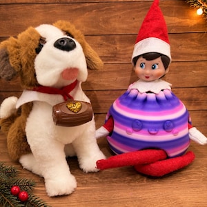 May include: A plush Saint Bernard dog with brown and white fur wearing a red collar with a brown barrel charm featuring a gold heart, positioned next to a small elf doll wearing a red hat and a round, purple and pink striped plastic shell.