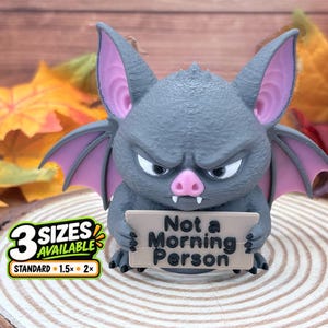 May include: A gray bat figurine with large pointed ears, pink inner ears, and a pink nose. The bat has a grumpy expression and holds a tan rectangular sign that reads "Not a Morning Person" in black text. A graphic indicates 3 sizes available.