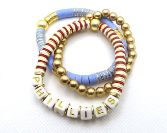 May include: Three bracelets stacked on top of each other. The top bracelet is light blue with gold accents. The middle bracelet is red and white striped with gold accents. The bottom bracelet is white with gold letters spelling out "PHILLIES".