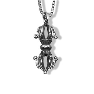 May include: A silver-toned metal pendant shaped like a vajra, featuring intricate symmetrical carvings and a central rounded band. The pendant hangs from a silver-toned box chain necklace, showcasing a polished finish with darkened recessed details.