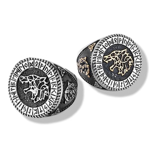 Puede incluir: Dos anillos plateados con un diseño de cabeza de lobo. Los anillos tienen un fondo oscuro con una cabeza de lobo dorada en el centro, rodeada por un borde circular con símbolos grabados. Los lados de los anillos también presentan detalles de cabeza de lobo.