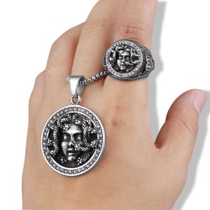 May include: A silver-toned metal ring and a matching circular pendant connected by a chain. Both pieces feature a detailed relief of a face with snakes for hair, accented with small clear stones along the circular borders of the jewelry.
