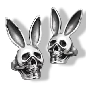 May include: Two silver rings, each featuring a skull design with long rabbit ears. The rings have a detailed, gothic aesthetic, with the skull's teeth and facial features clearly visible. The rings are set against a white background.
