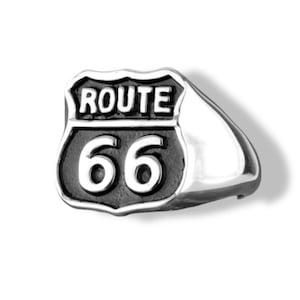 May include: A silver ring featuring the iconic Route 66 sign. The ring has a polished finish with the words "ROUTE" and the number "66" in a bold, black font. The design is a classic symbol of American road trips.