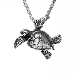 May include: A silver-toned metal pendant shaped like a sea turtle, featuring detailed textured flippers and a smooth, segmented shell design. The pendant is suspended from a matching silver-toned metal box chain necklace with a polished finish.