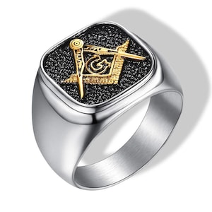 Puede incluir: Anillo de acero inoxidable con una cara cuadrada que presenta un símbolo masónico dorado sobre un fondo negro texturizado. El anillo tiene un acabado pulido y reflectante.