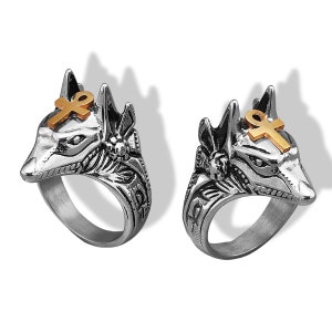 May include: Two silver-toned Anubis rings with gold-colored ankh symbols. The rings feature detailed carvings of the jackal-headed deity, showcasing intricate designs and a polished finish. The rings are displayed on a white background.