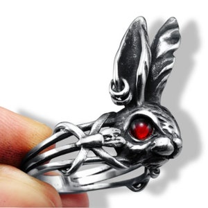 May include: A silver-colored ring featuring a detailed rabbit head with a red gemstone eye. The rabbit's ears have a hoop earring, and the ring has a unique, intertwined design around the band. The ring is held by a hand.